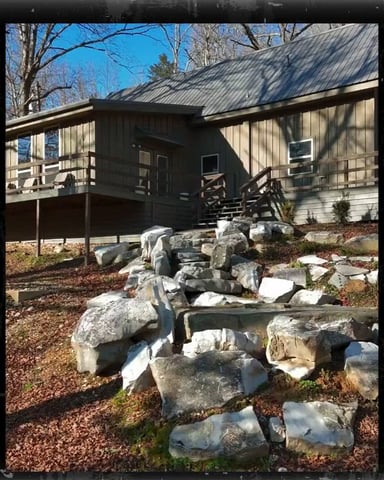 Modern cabin with metal roof and deck, surrounded by landscaping rocks and bare trees on clear day