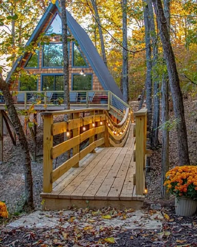 Modern A-frame treehouse with large windows and deck nestled among autumn trees, wooden bridge entrance with string lights and orange mums.
