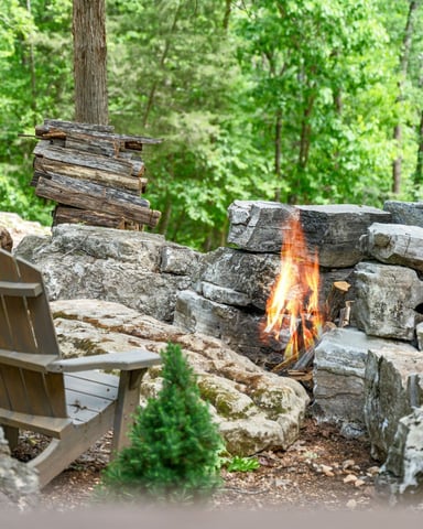 Stone fire pit with active flames surrounded by Adirondack chairs in a wooded forest setting