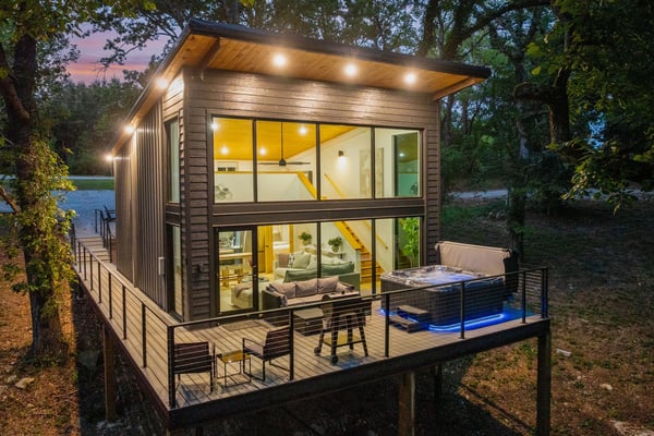 Modern elevated cabin with large windows, lit interior, wooden deck with seating and hot tub, surrounded by trees and water at dusk