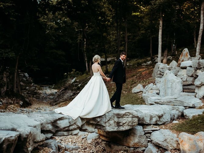 Bride and groom holding hands on rocky terrain surrounded by forest and stone ruins.